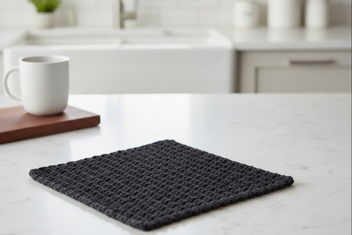 A black crocheted dish cloth on a marble surface with a coffee mug on a wood tray and a sink in the background