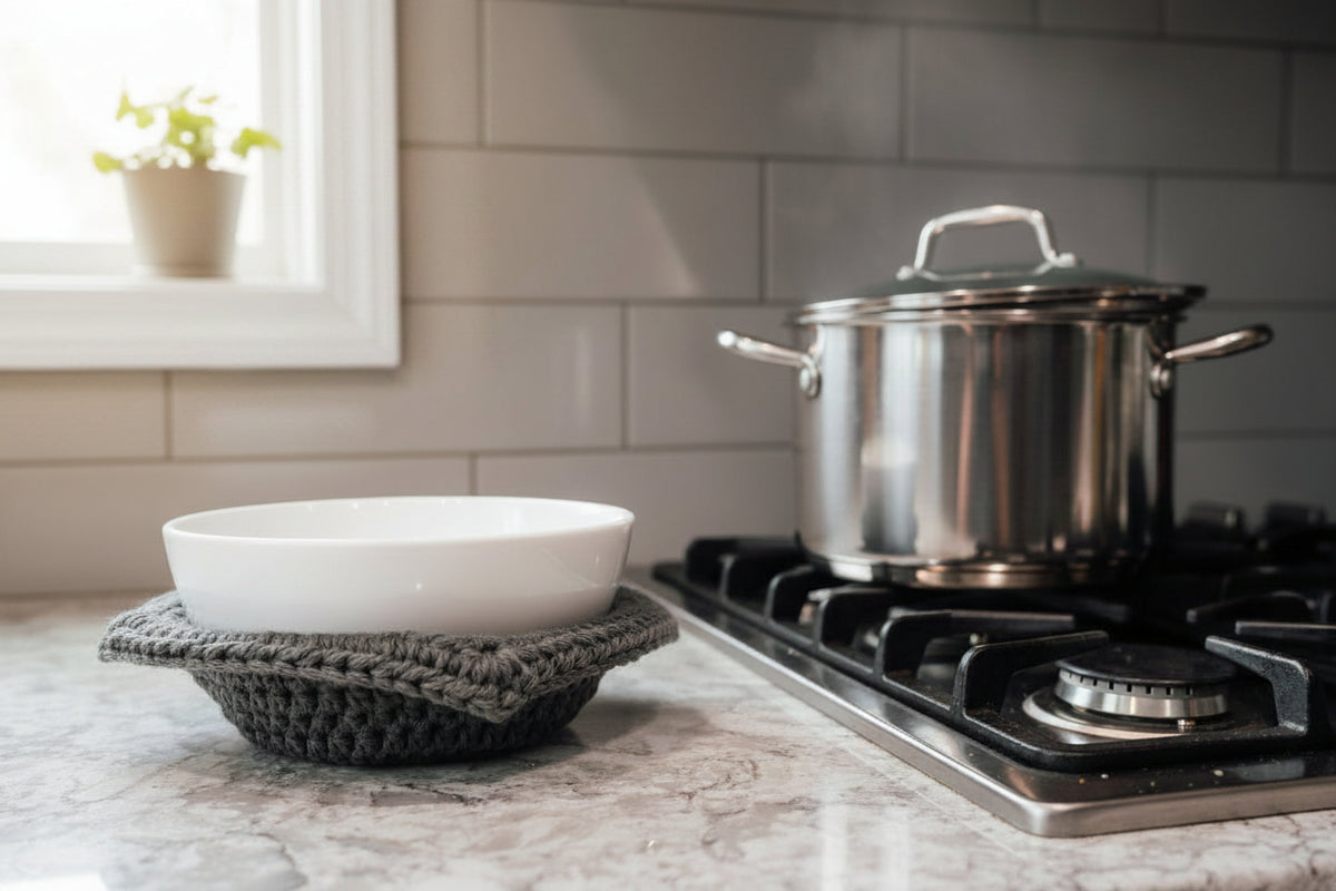 A white bowl in a grey crocheted bowl cozy next to a pot on a stove with a window in the background