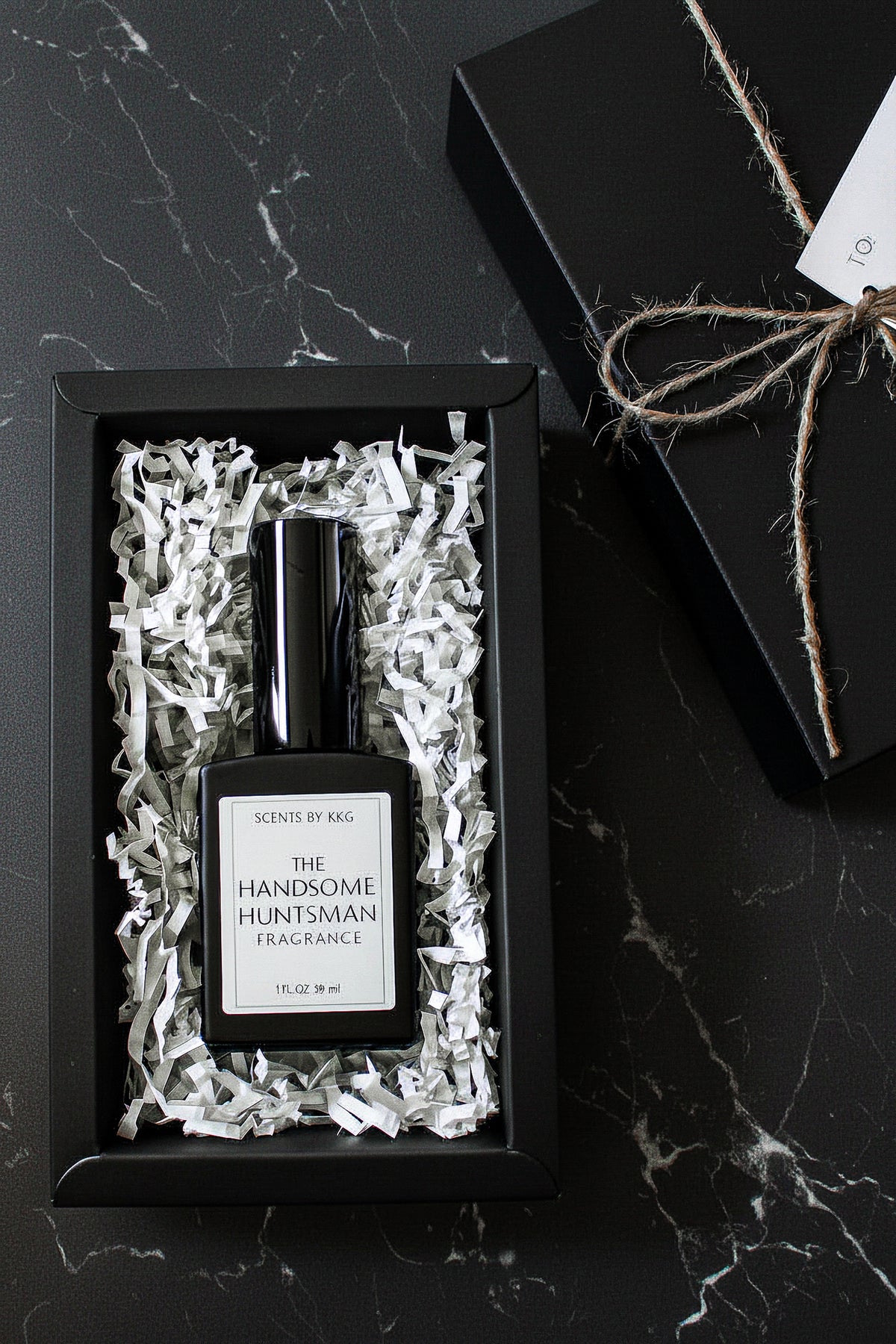 A black rectangular fragrance bottle labeled 'The Handsome Huntsman Fragrance' by Scents by K&G, presented in white crinkle paper inside a black gift box on a dark marble surface, with the box top and twine visible beside it.