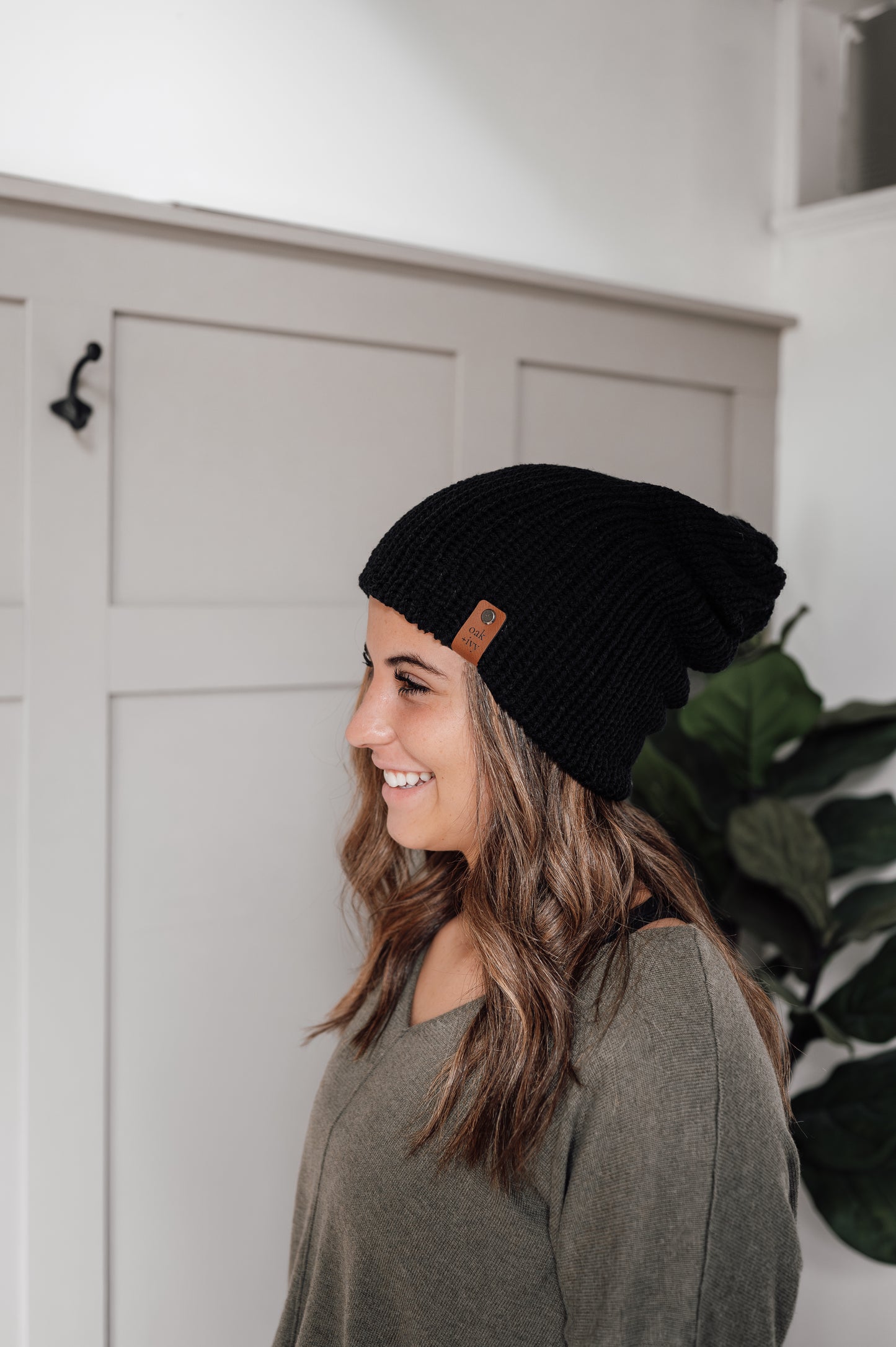 A woman wearing a black knit beanie with a visible brand tag, standing in a room with a white door and a potted plant in the background.
