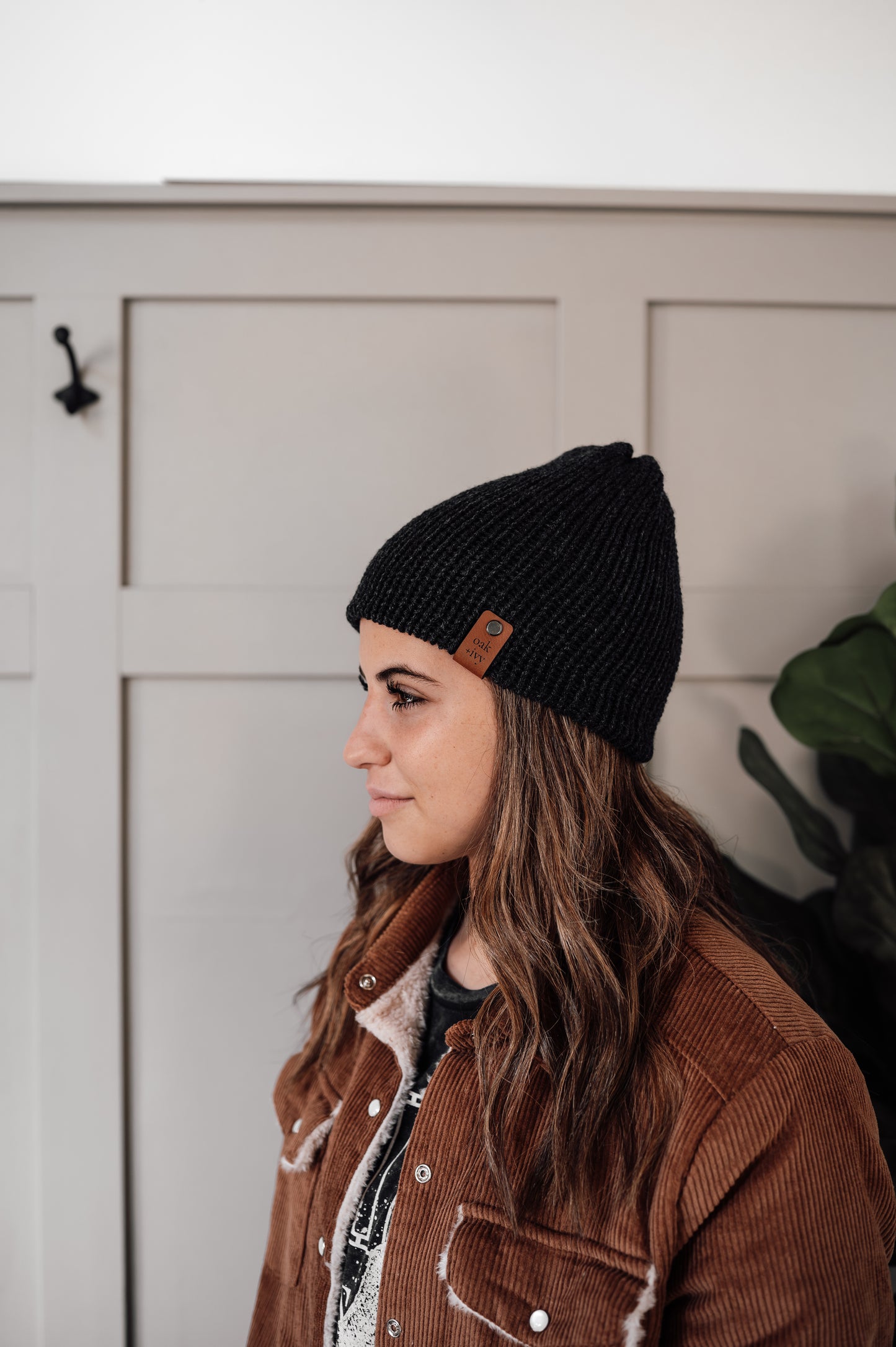 A person wearing a dark grey heather knit beanie with a visible brand tag on the brim, standing indoors.