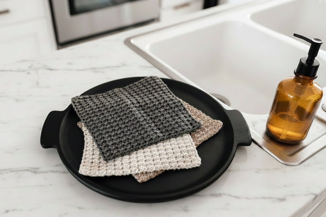 A set of three neutral crocheted dish cloths on a black tray next to a sink and a soap dispenser