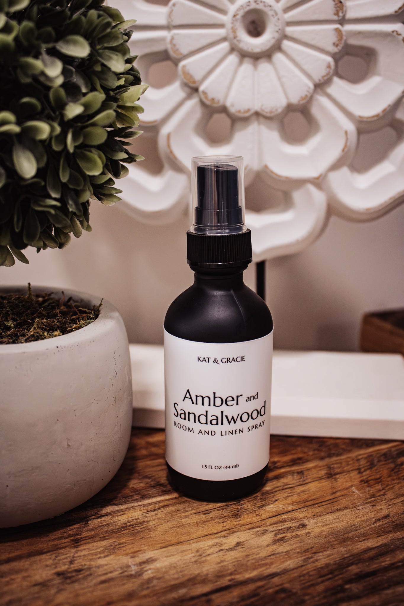 Room spray bottle labeled 'Amber and Sandalwood' by Kat & Gracie on a wooden surface with decorative elements.
