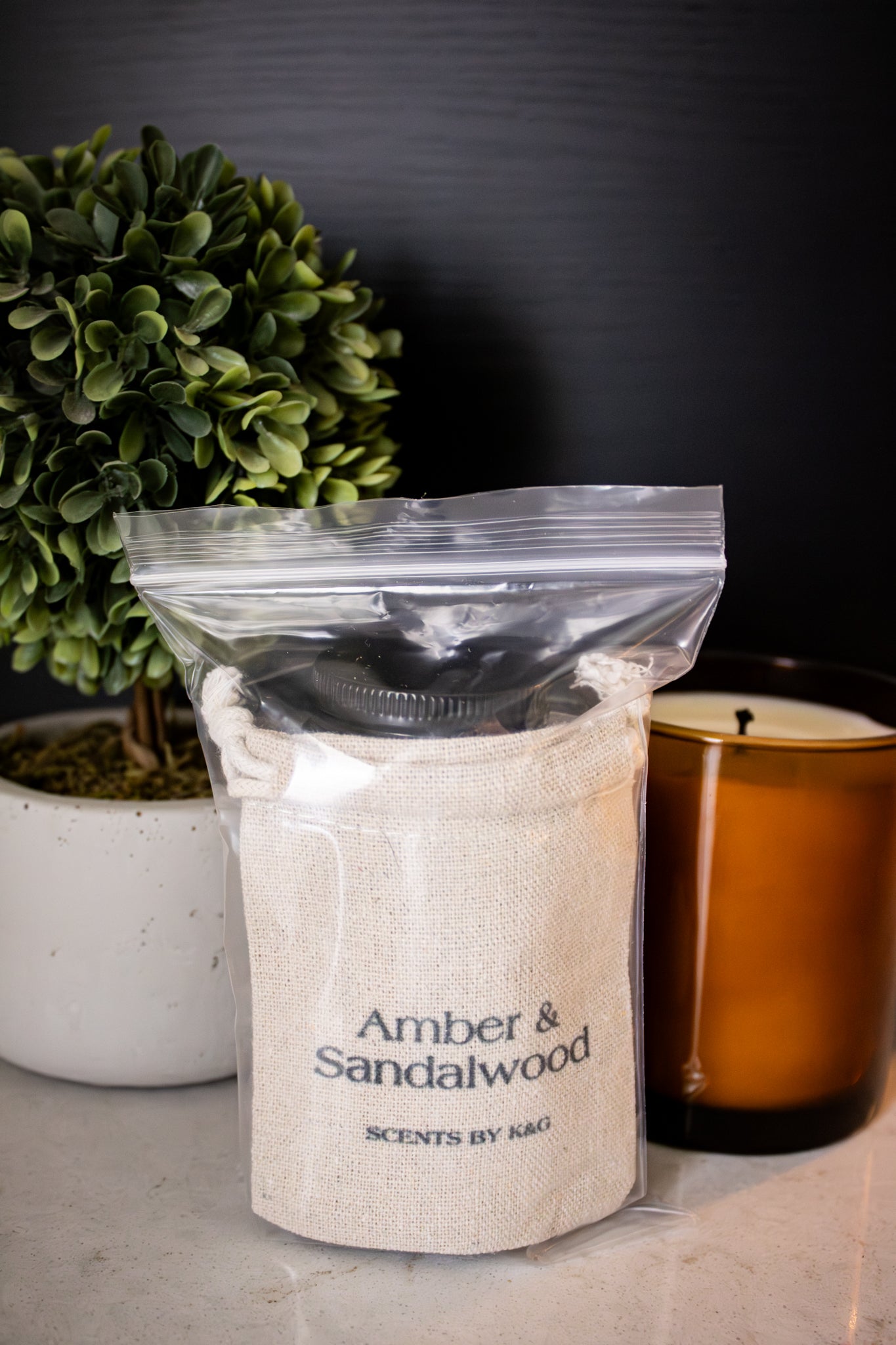 Package of 'Amber & Sandalwood' scented product on a surface with a plant and candle in the background.