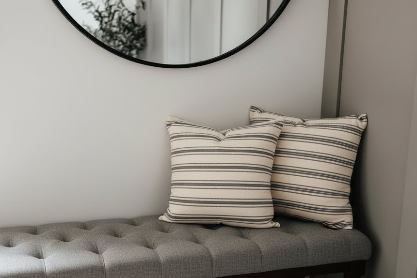 Two square throw pillows with horizontal black and beige stripes resting on a gray tufted bench against a light gray wall, with a round black-framed mirror above.