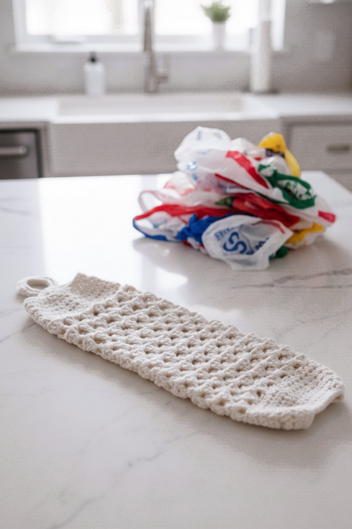 A handmade off-white crochet bag saver with a hanging loop at the top rests on a white marble kitchen countertop, with a pile of colorful plastic shopping bags in the blurred background near a sink.