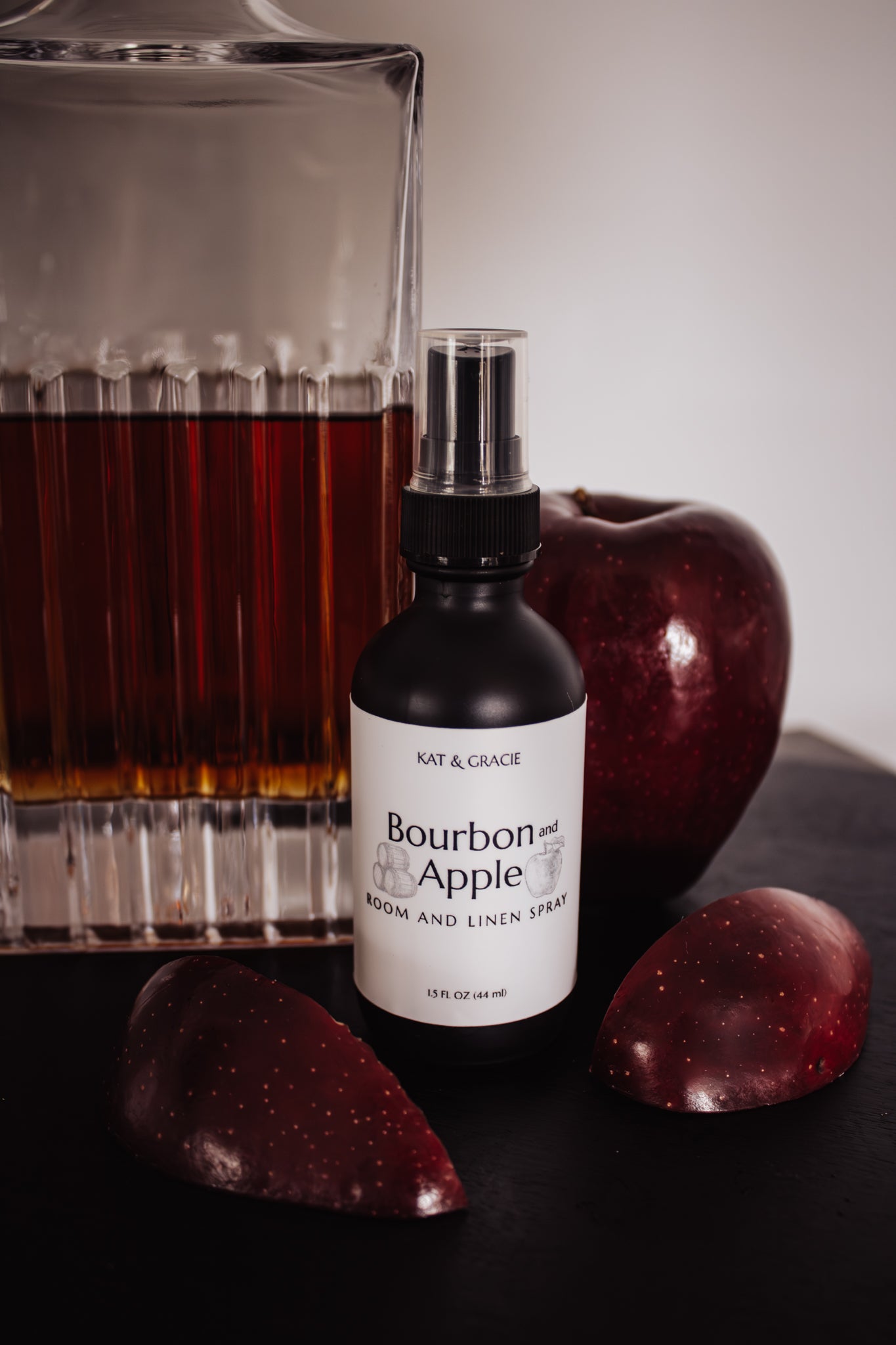 Bourbon and Apple room and linen spray bottle with apples and a glass of bourbon on a dark surface.