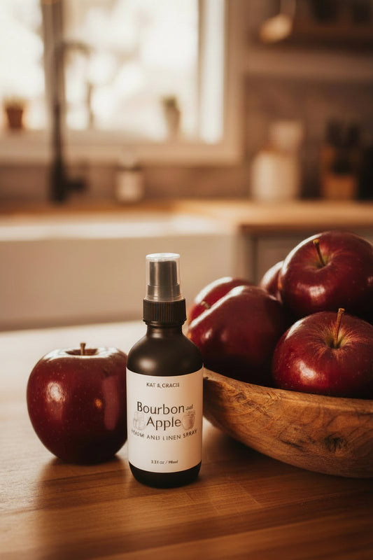 Bourbon and Apple room and linen spray by Kat & Gracie on a wooden surface next to a bowl of red apples in a kitchen