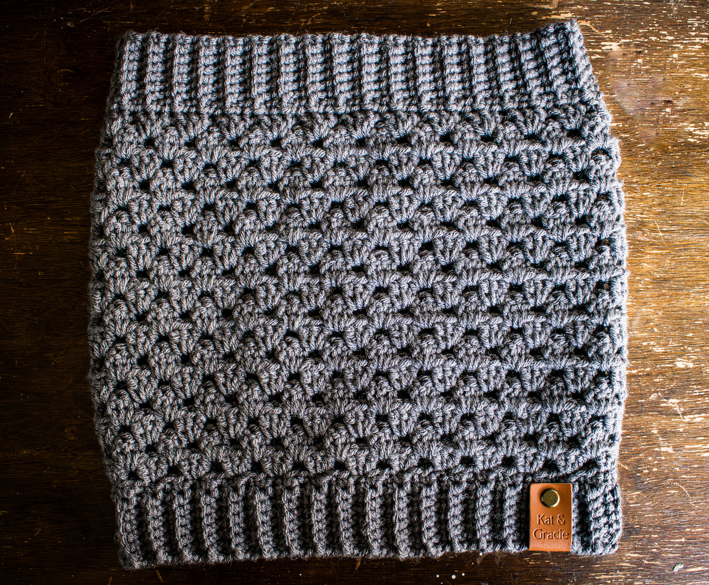Gray crocheted cowl with a brand tag on a wooden surface