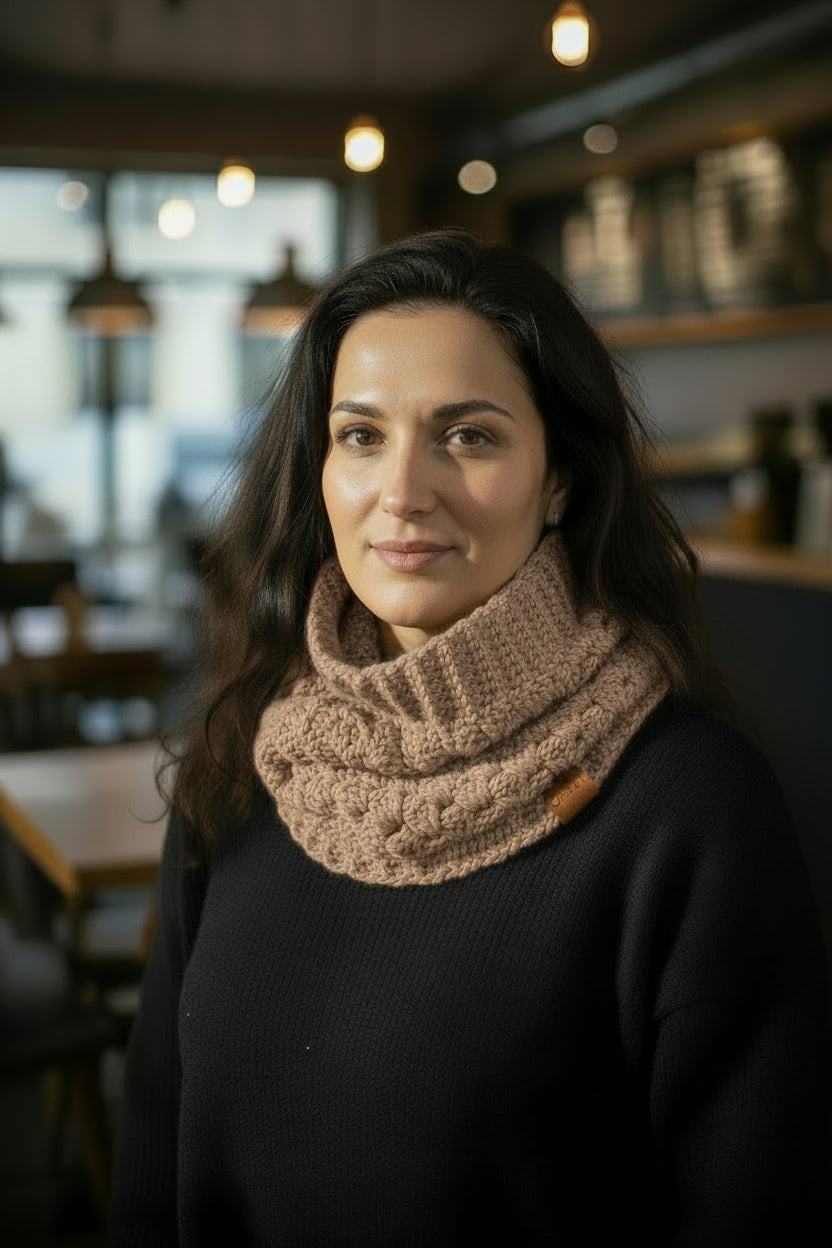 A woman with long brown hair wearing a crocheted beige cowl neck warmer and a black sweater, standing in an indoor setting.