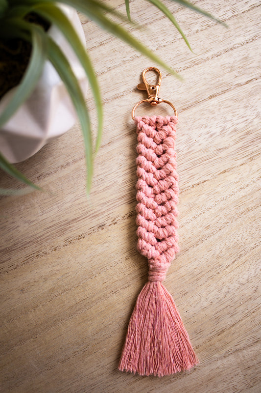 Pink braided tassel keychain on a wooden surface with a plant in the corner.