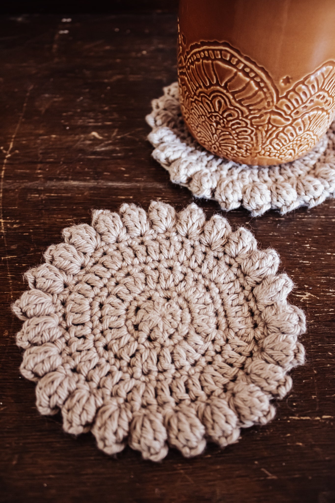 Two beige crocheted coasters on a wooden surface with a ceramic mug.