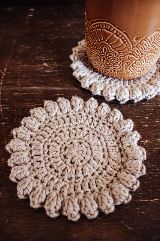 Two beige crocheted coasters on a wooden surface with a ceramic mug.