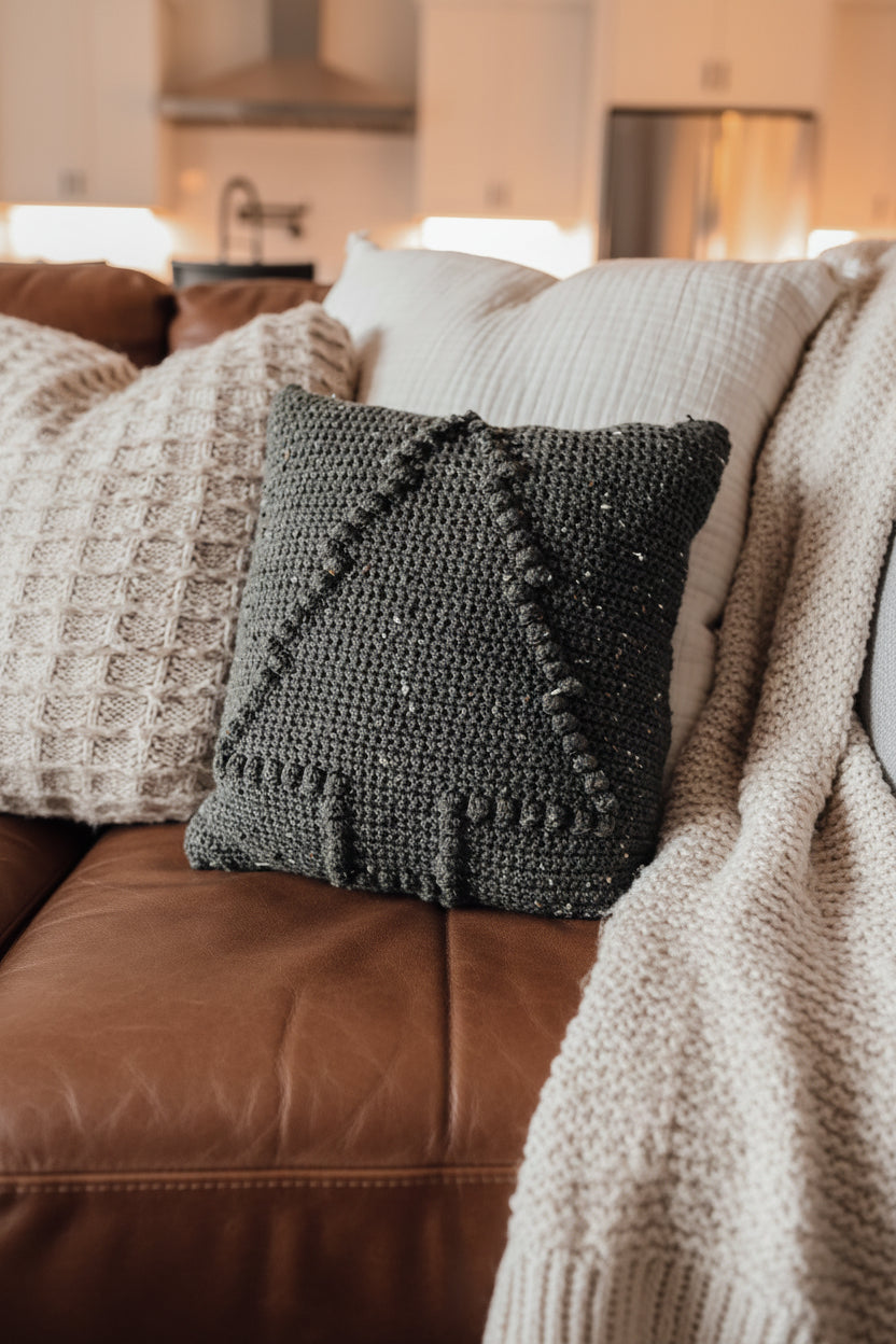 A crocheted holiday pillow with a dark green tweed pattern, featuring a bobble tree design, placed on a sofa with other decorative pillows.
