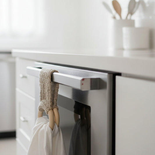 A beige crocheted towel holder with wooden loops holding a white cloth, hanging on a stainless steel oven handle in a bright, modern kitchen.