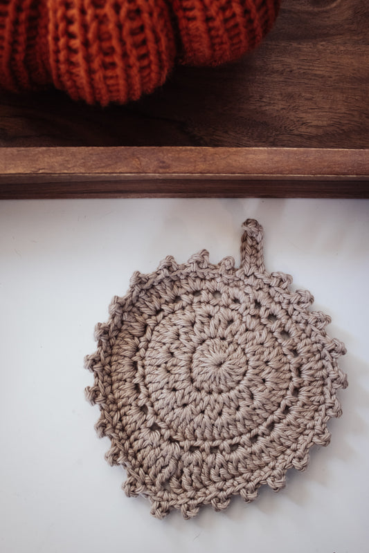 Khaki crocheted leaf coaster on a white surface with an orange pumpkin in the background