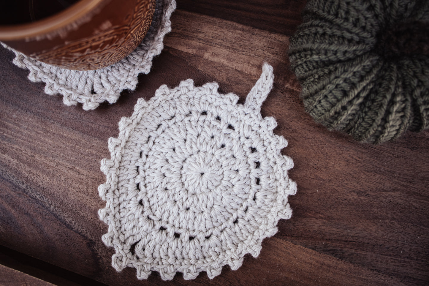 Crocheted off-white coaster on a wooden surface with an olive green knitted pumpkin and amber-colored ceramic mug