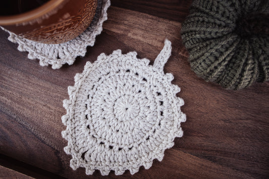 Crocheted off-white coaster on a wooden surface with an olive green knitted pumpkin and amber-colored ceramic mug