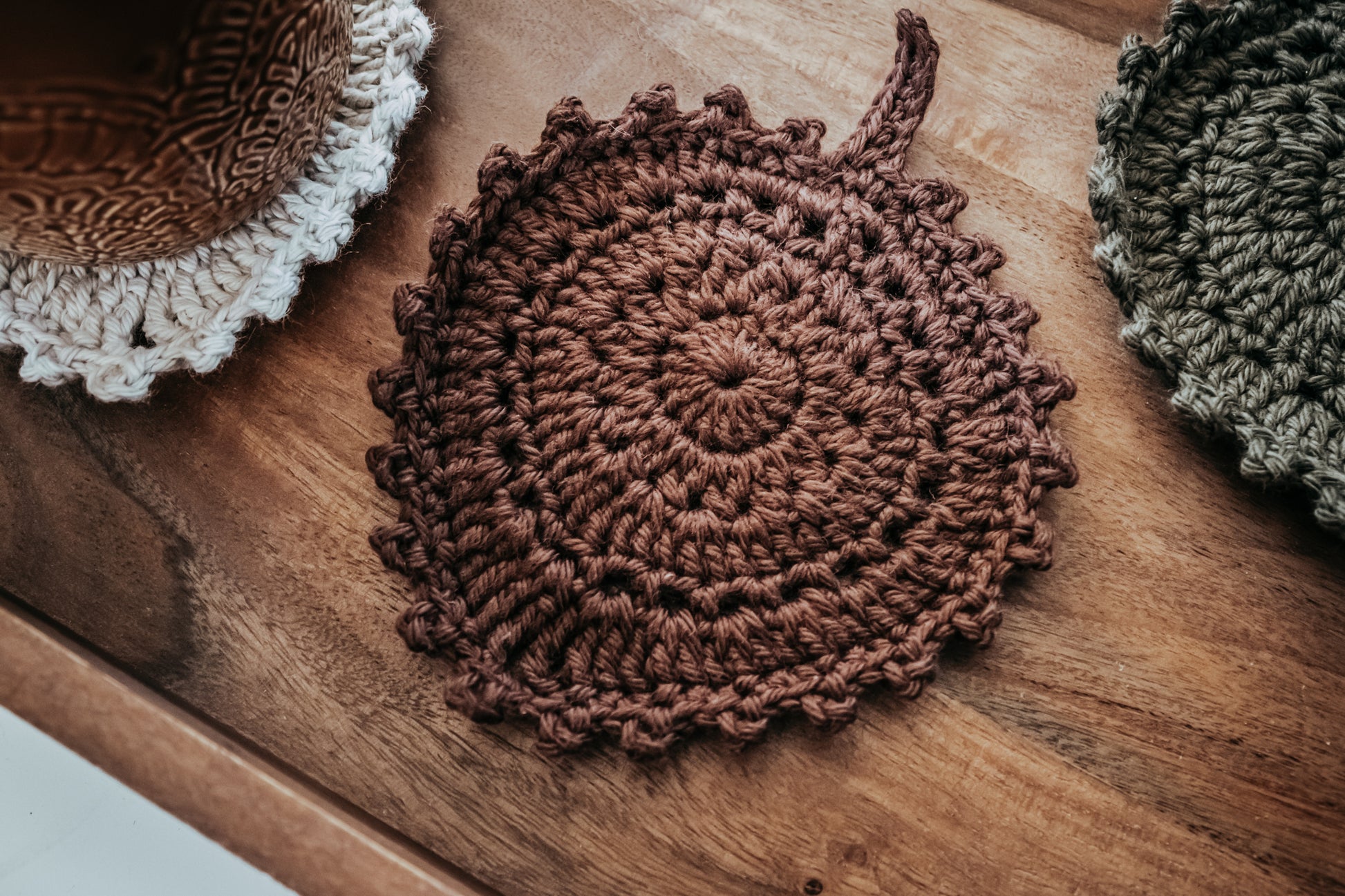 Crocheted leaf coasters in brown, off-white, and olive green on a wooden tray.
