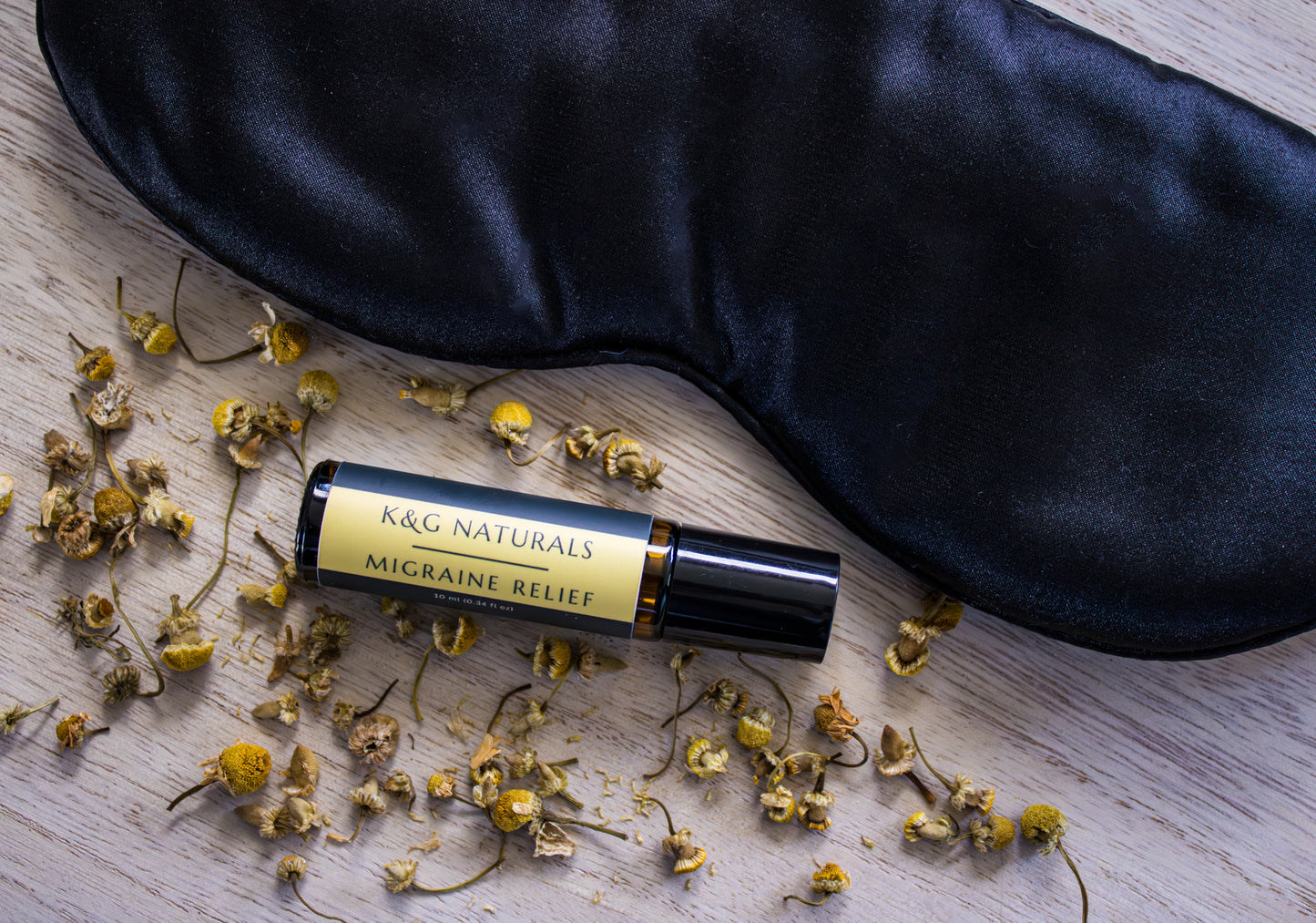 K&G Naturals Migraine Relief rollerball with dried chamomile and a black sleep mask on a wooden surface