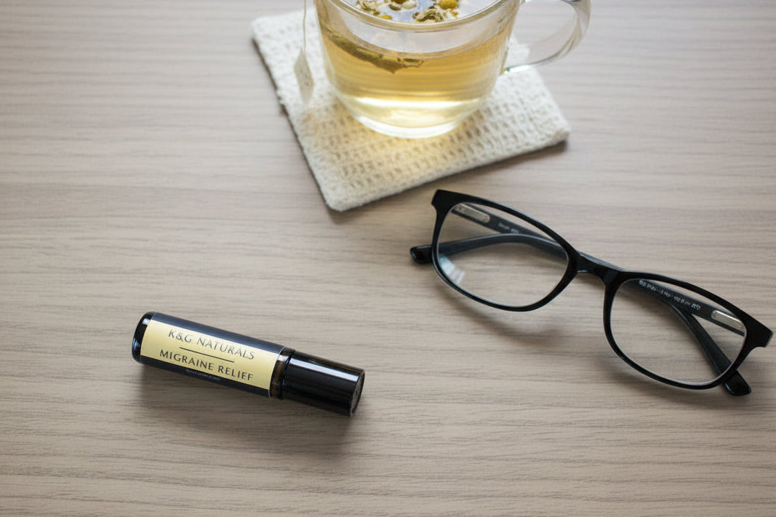 A bottle of K&G Naturals Migraine Relief rollerball with a black cap and gold label, next to black eyeglasses and a cup of tea on a light surface
