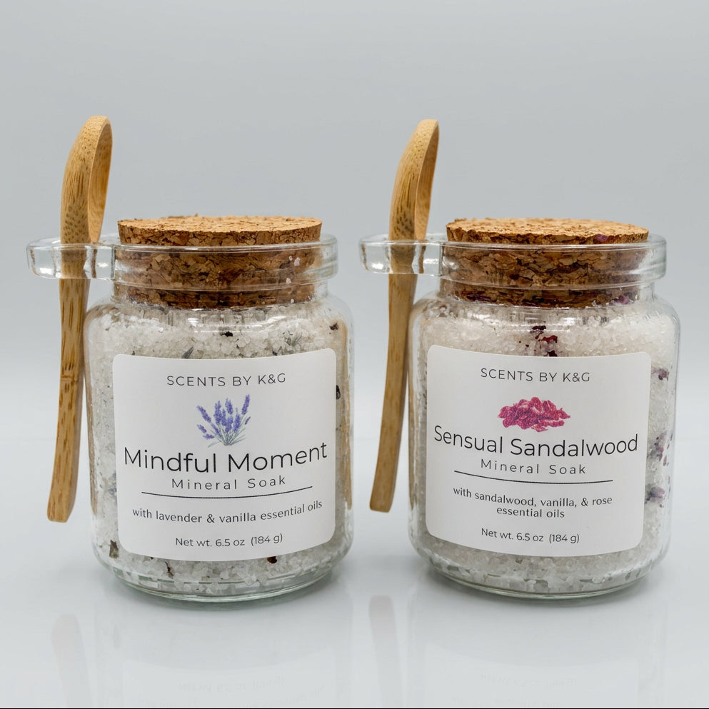 Two glass jars of mineral soak bath salts with cork lids and wooden spoon handles, labeled 'Scents by K&G' Mindful Moment and Sensual Sandalwood, on a reflective white surface.