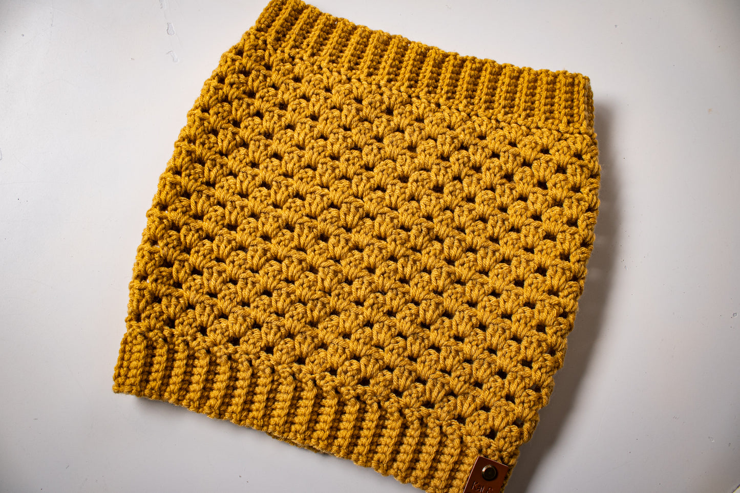 Mustard colored crocheted cowl with brand tag on a white background.