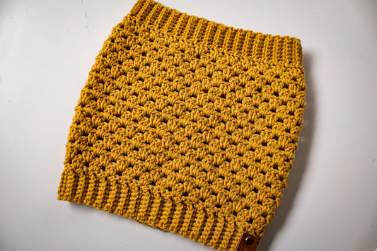 Mustard colored crocheted cowl with brand tag on a white background.