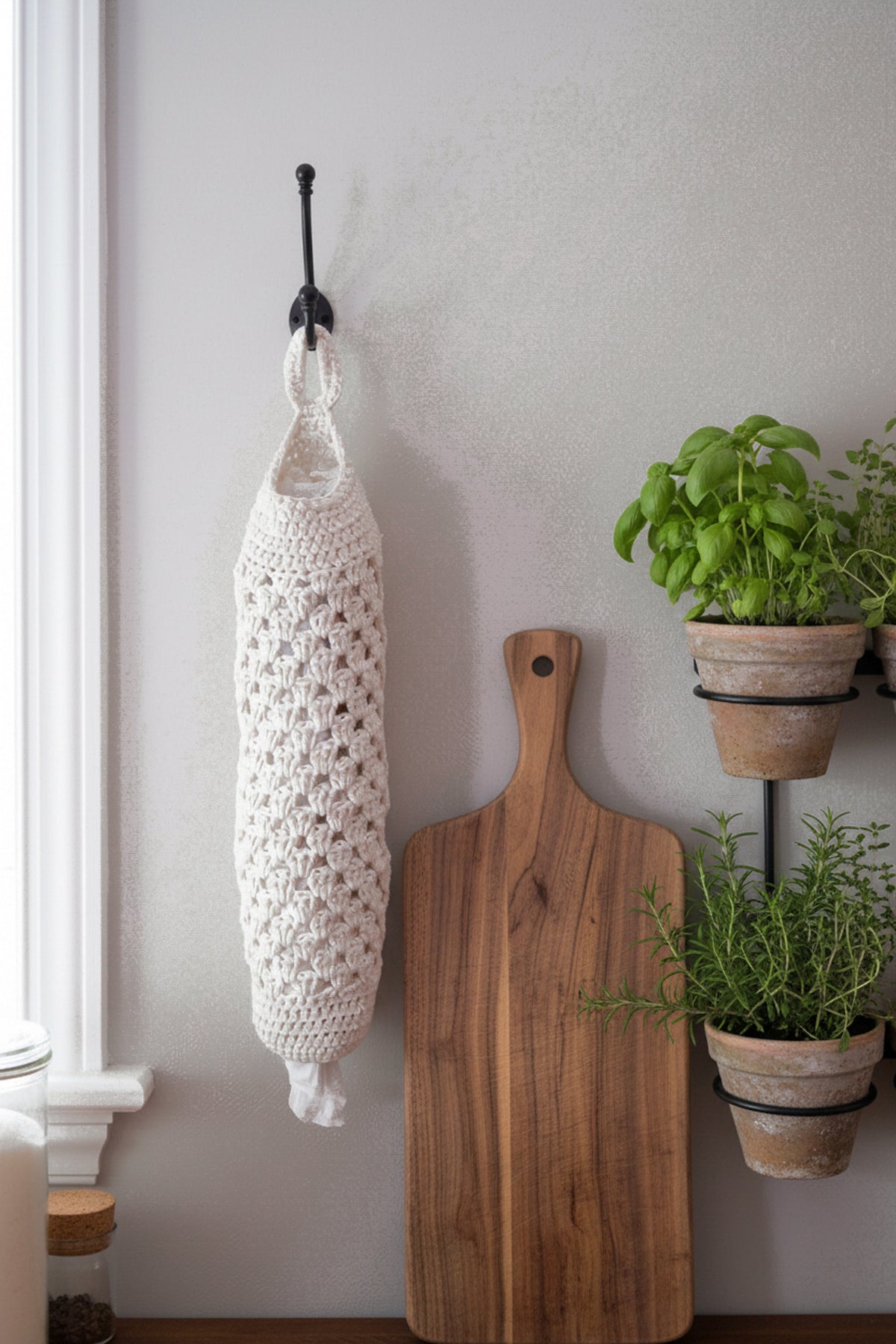A white crocheted hanging plastic bag saver with a top loop hangs from a black wall hook on a light gray wall, styled next to a wooden cutting board and potted green plants.