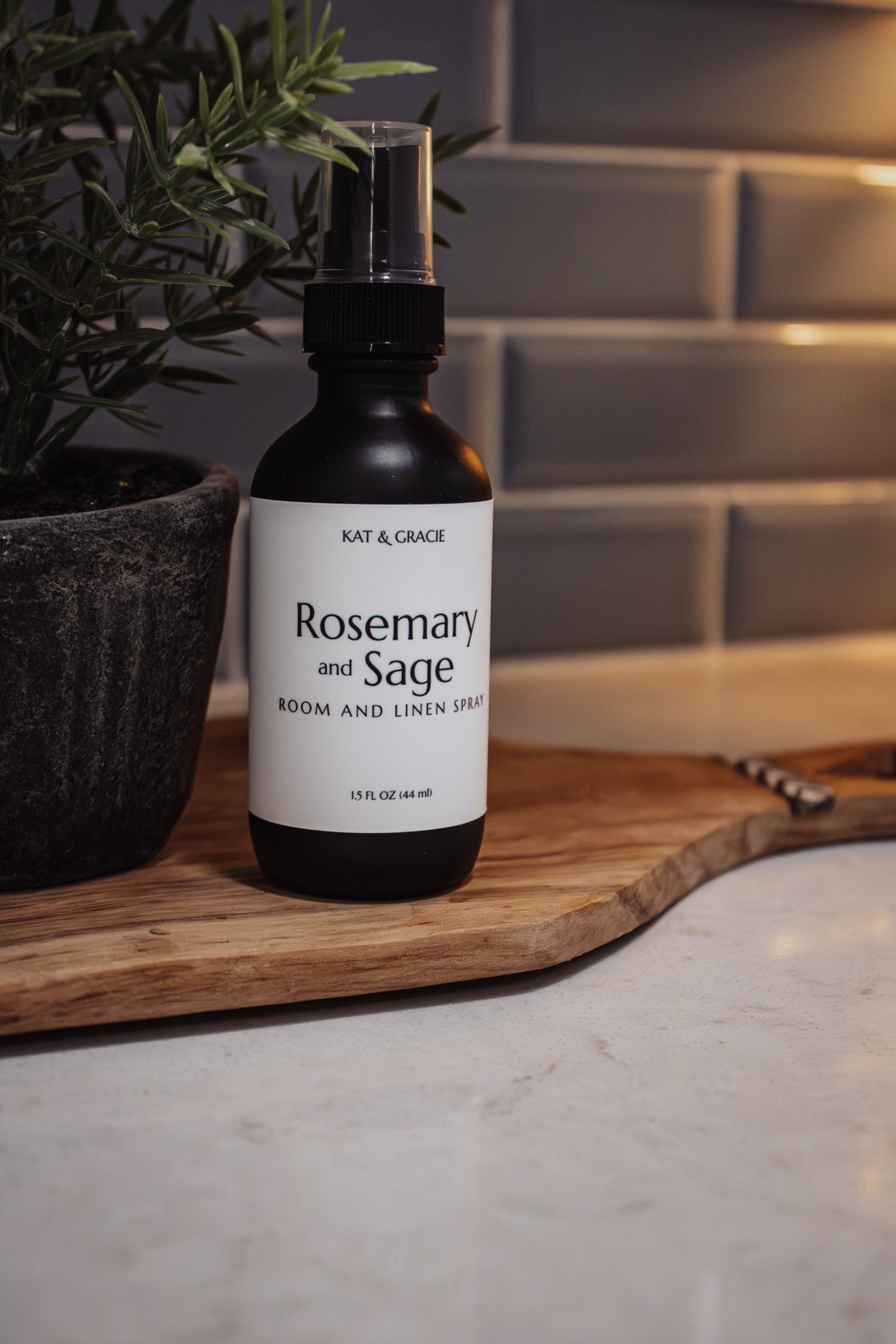 Bottle of Kat & Gracie Rosemary and Sage Room and Linen Spray on a wooden cutting board with a plant in the background.