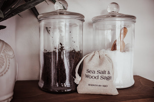 Two glass jars with lids and a small drawstring bag labeled 'Sea Salt & Wood Sage' on a wooden surface.