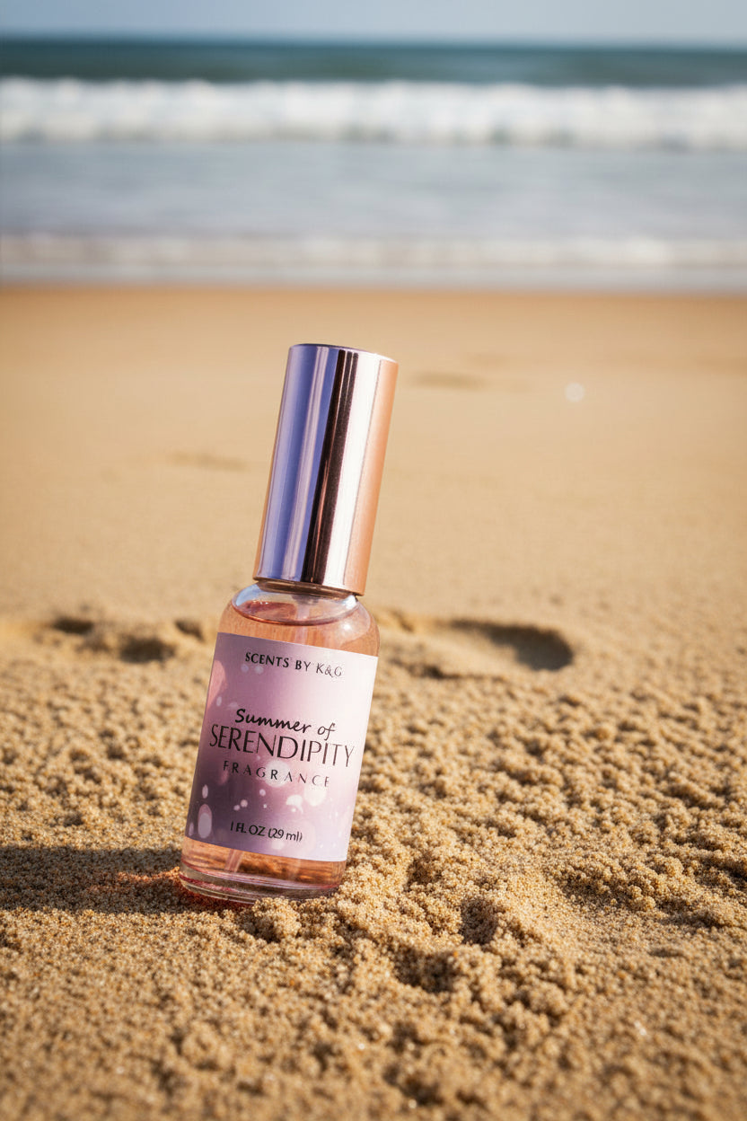 A bottle of Scents by K&G 'Summer of Serendipity' fragrance, featuring a metallic rose gold cap and a pink label, set in the sand on the beach.