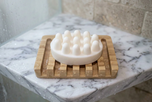 A shampoo bar featuring massager nodules sitting on a bamboo soap dish on a marble shelf in a shower.