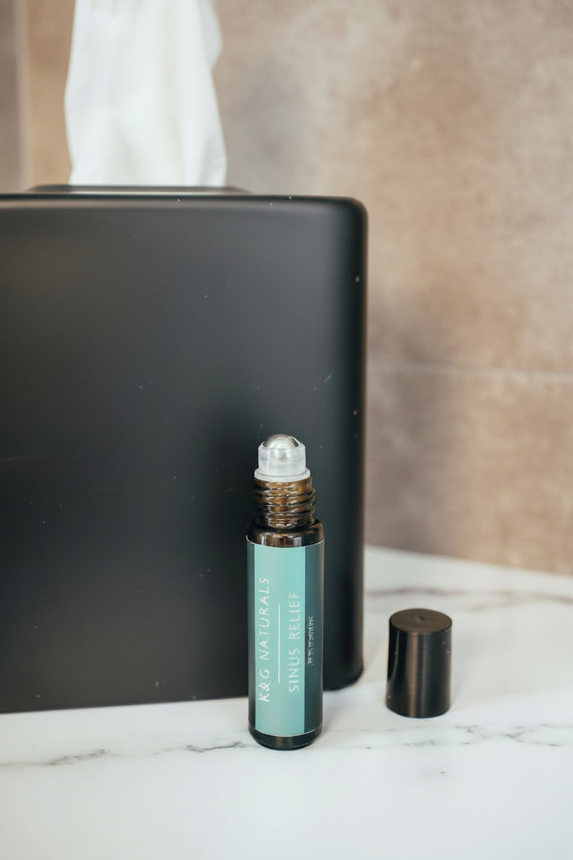 A small amber rollerball bottle labeled K&G Naturals Sinus Relief stands on a white surface in front of a black tissue box, with a black cap beside it