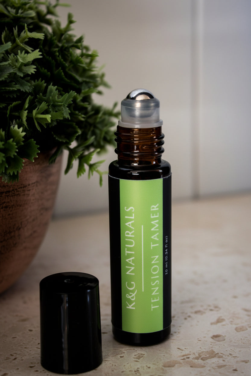 A small amber glass roller bottle with a bright green label reading 'K&G NATURALS TENSION TAMER' stands on a light marble surface, with its black cap placed beside it. The background is a softly blurred beige-toned interior wall.