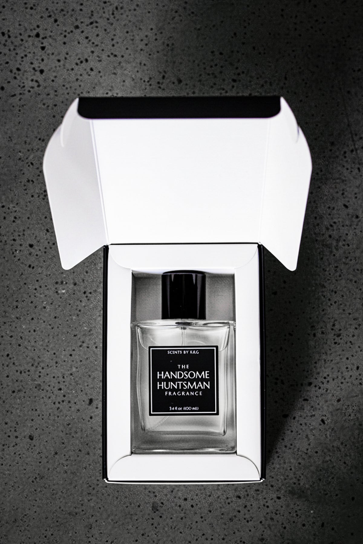 A clear glass perfume bottle with a black cap and black label reading 'The Handsome Huntsman Fragrance' by Scents by K&G, presented in an open black and white gift box on a dark textured surface.