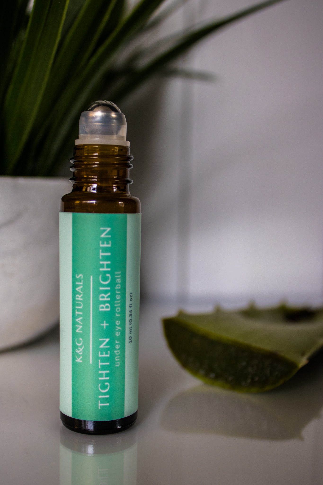 K&G Naturals under eye roller bottle with green label on a reflective surface with a plant in the background