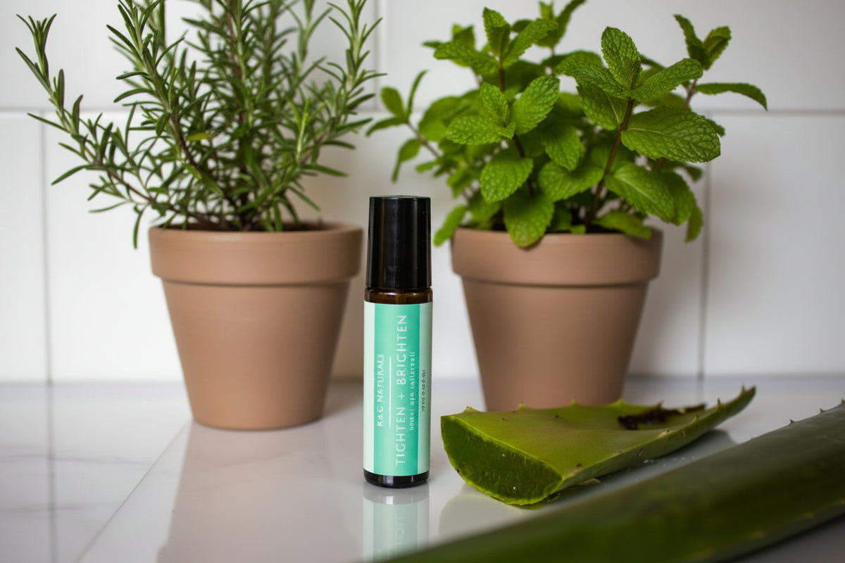 An amber rollerball labeled K&G Naturals Tighten + Brighten with a mint green label, standing on a white surface beside an aloe vera leaf, peppermint plant, and rosemary plant