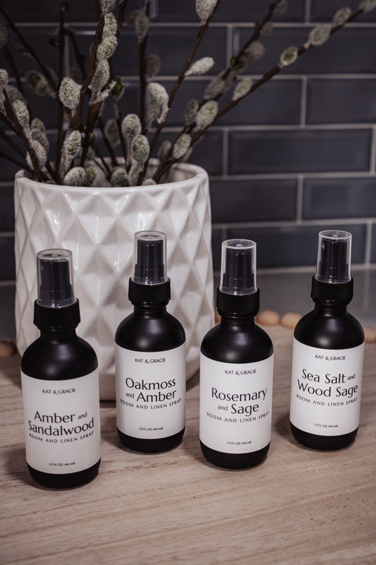 Four bottles of room sprays on a wooden surface with a vase of willow branches in the background.