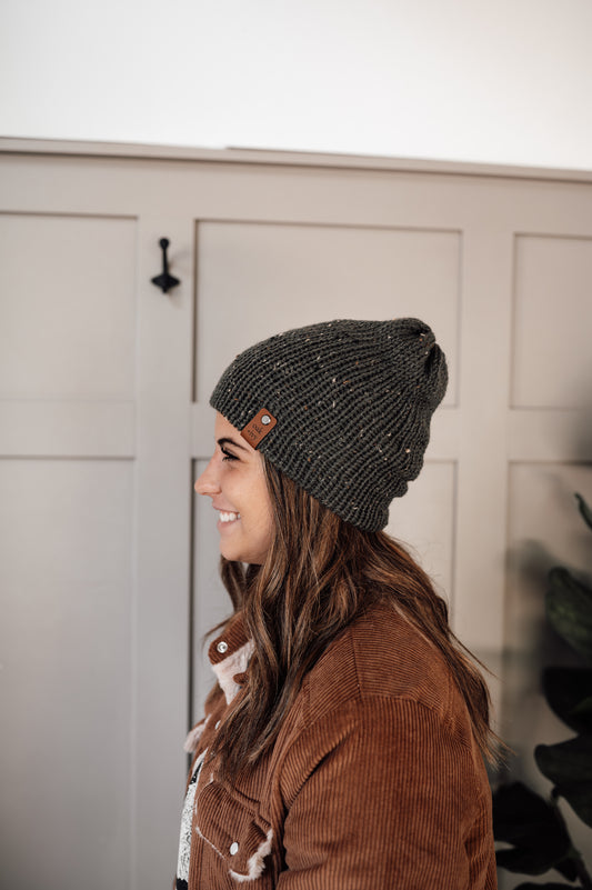 A person wearing a dark green tweed beanie with camel colored brand tag.